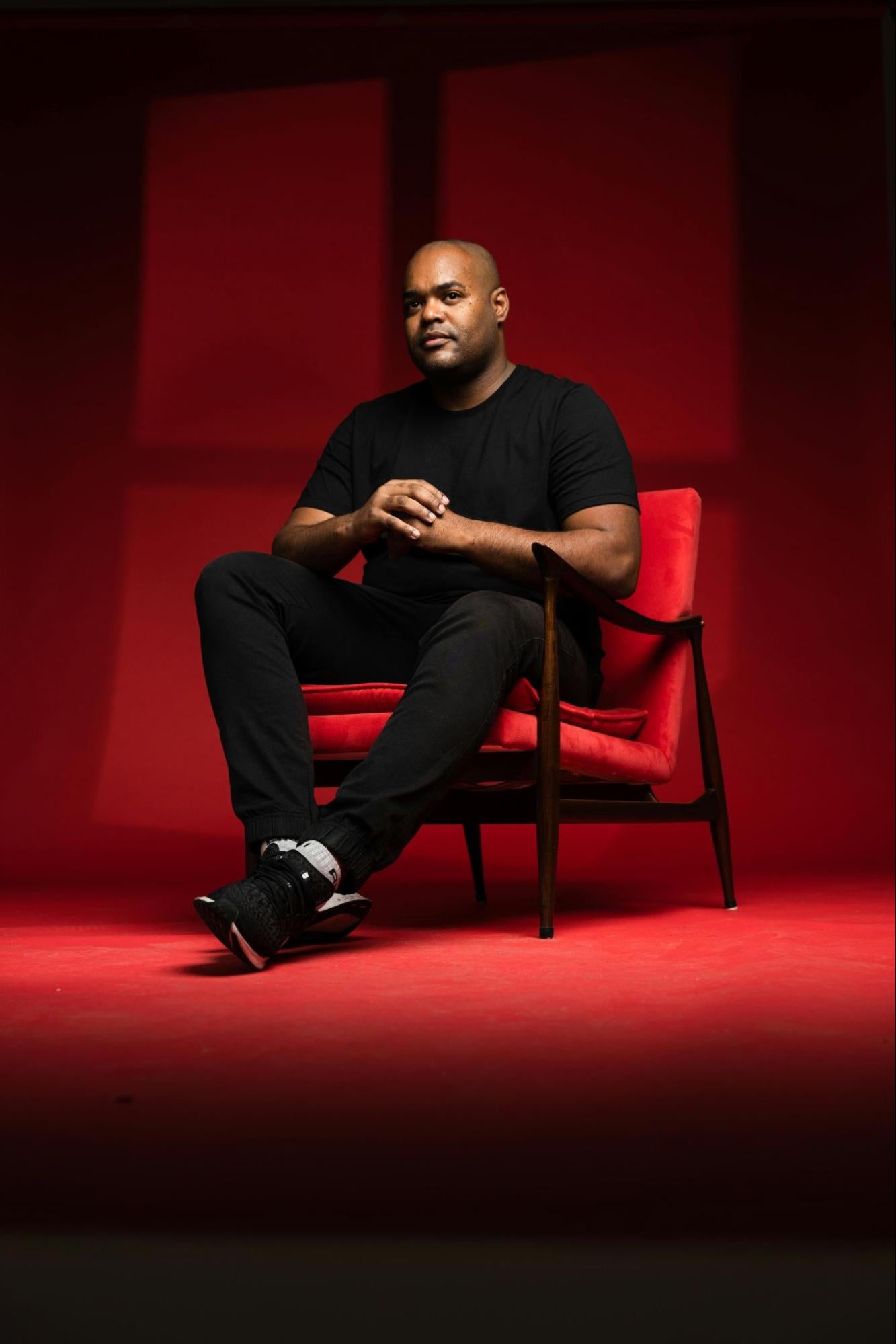 A man in a dark T-shirt sits on a red chair in a carpeted room.