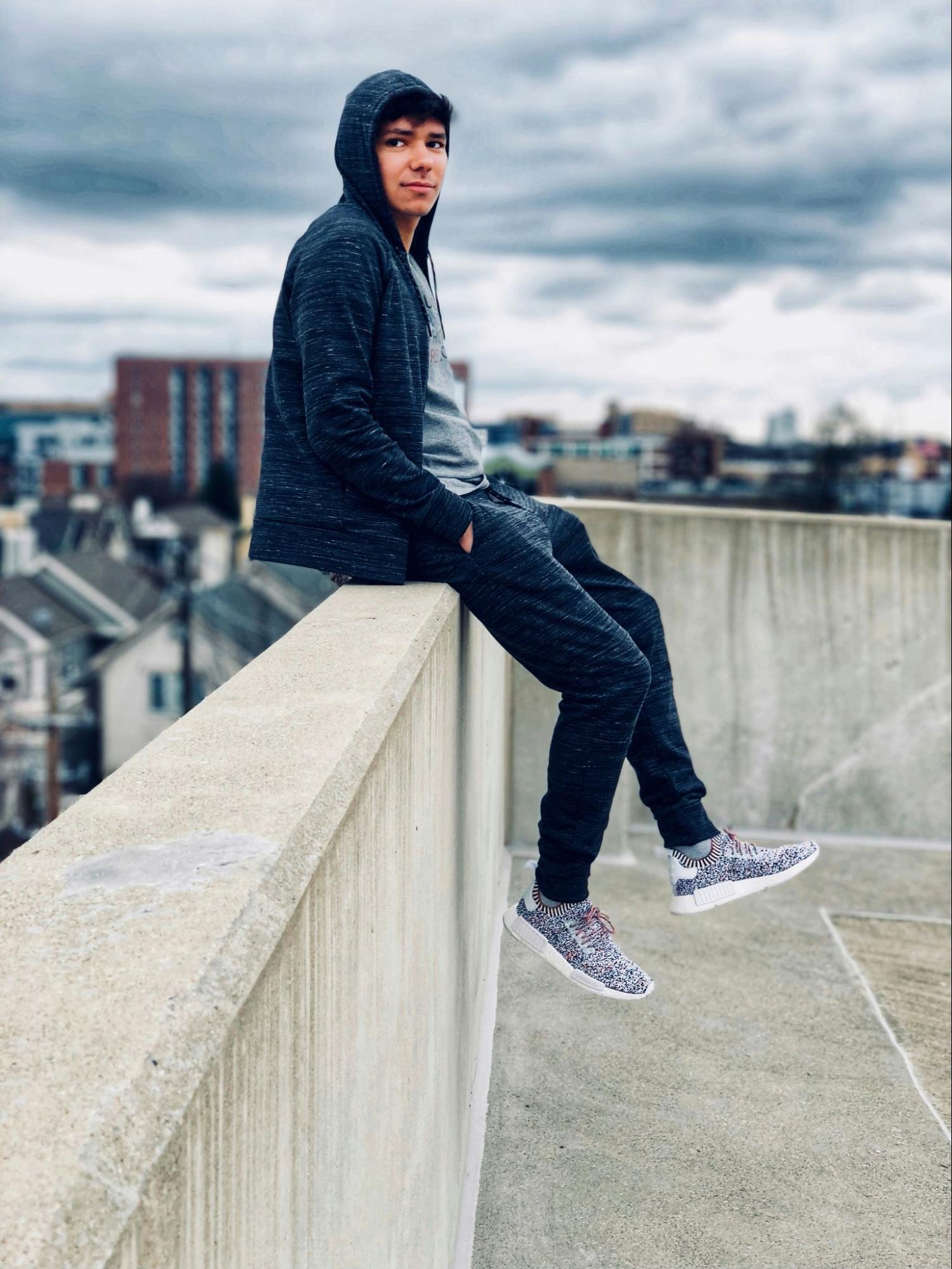 A man wearing joggers and a hoodie sits atop a concrete wall.