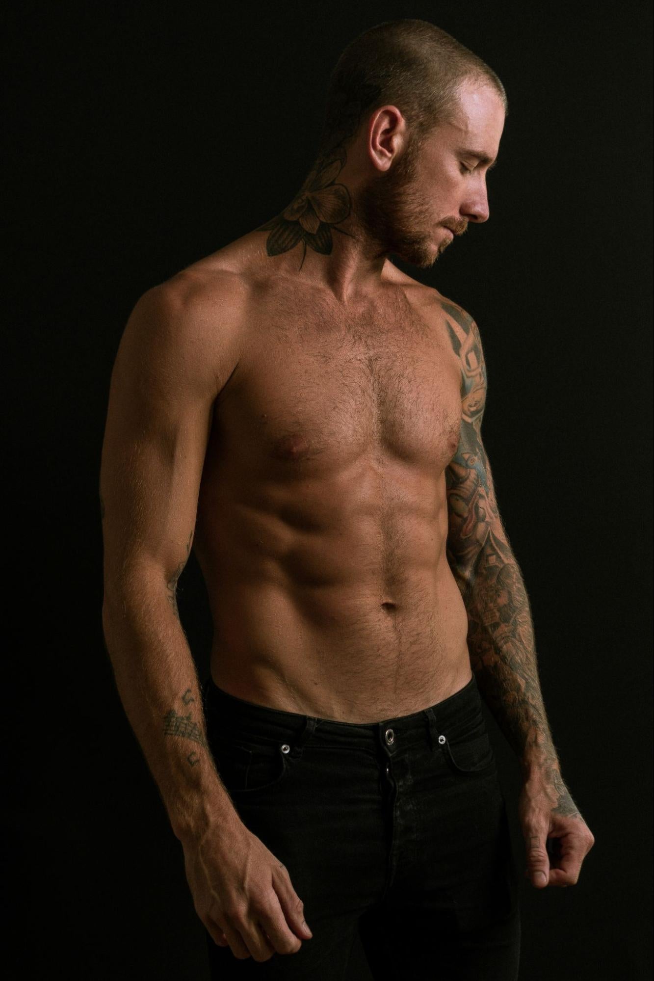 A tattooed man in his underwear stands in a dark room.