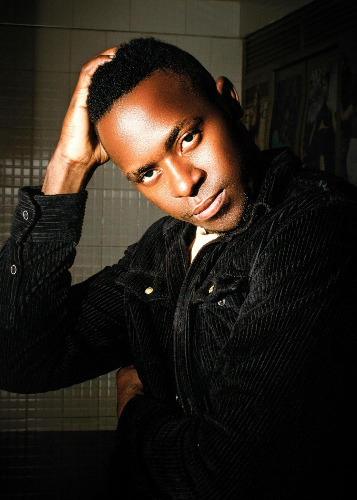 A man dressed in a dark corduroy jacket poses in a tiled room.