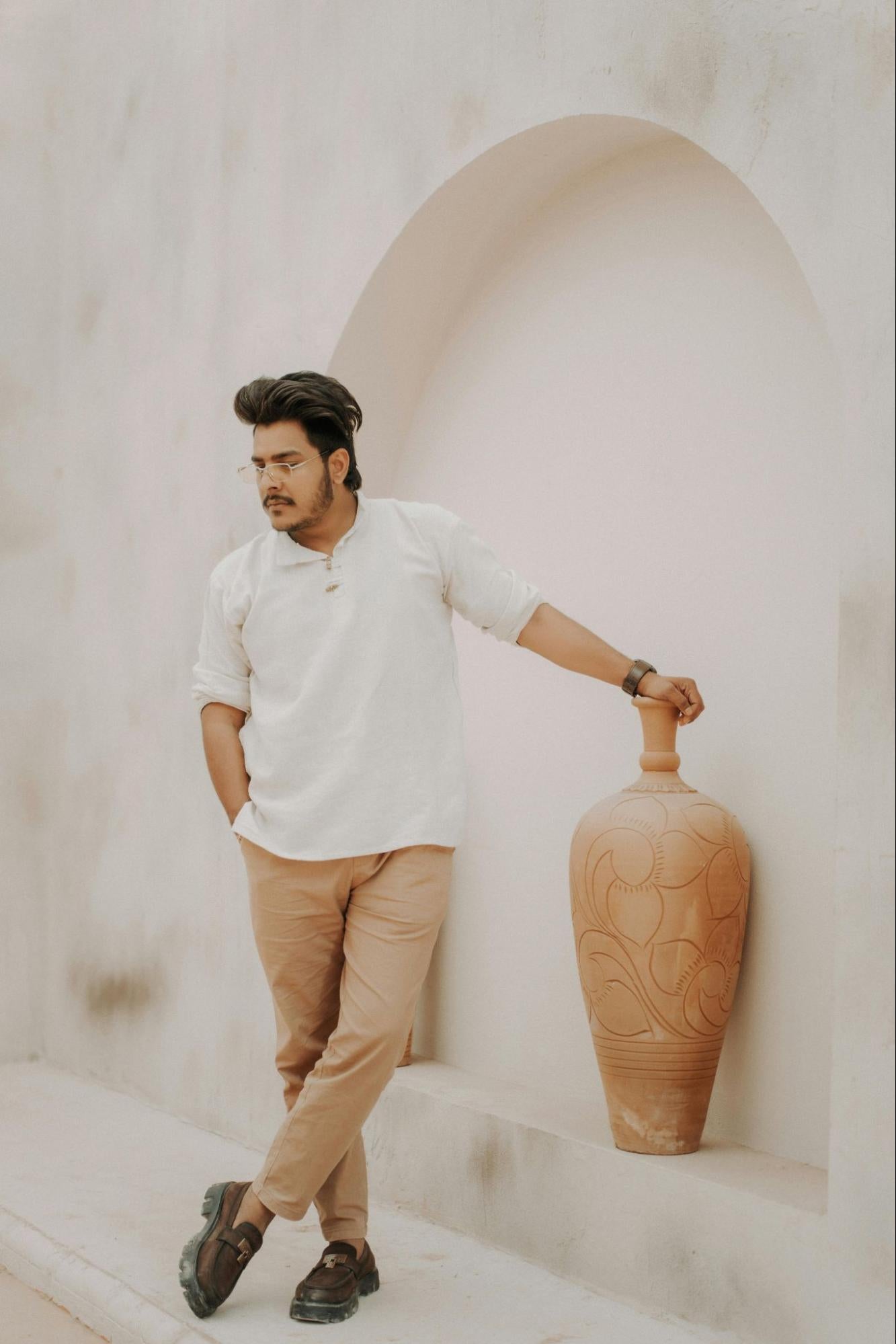 A stylish man wearing chinos stands next to a large embroidered vase.