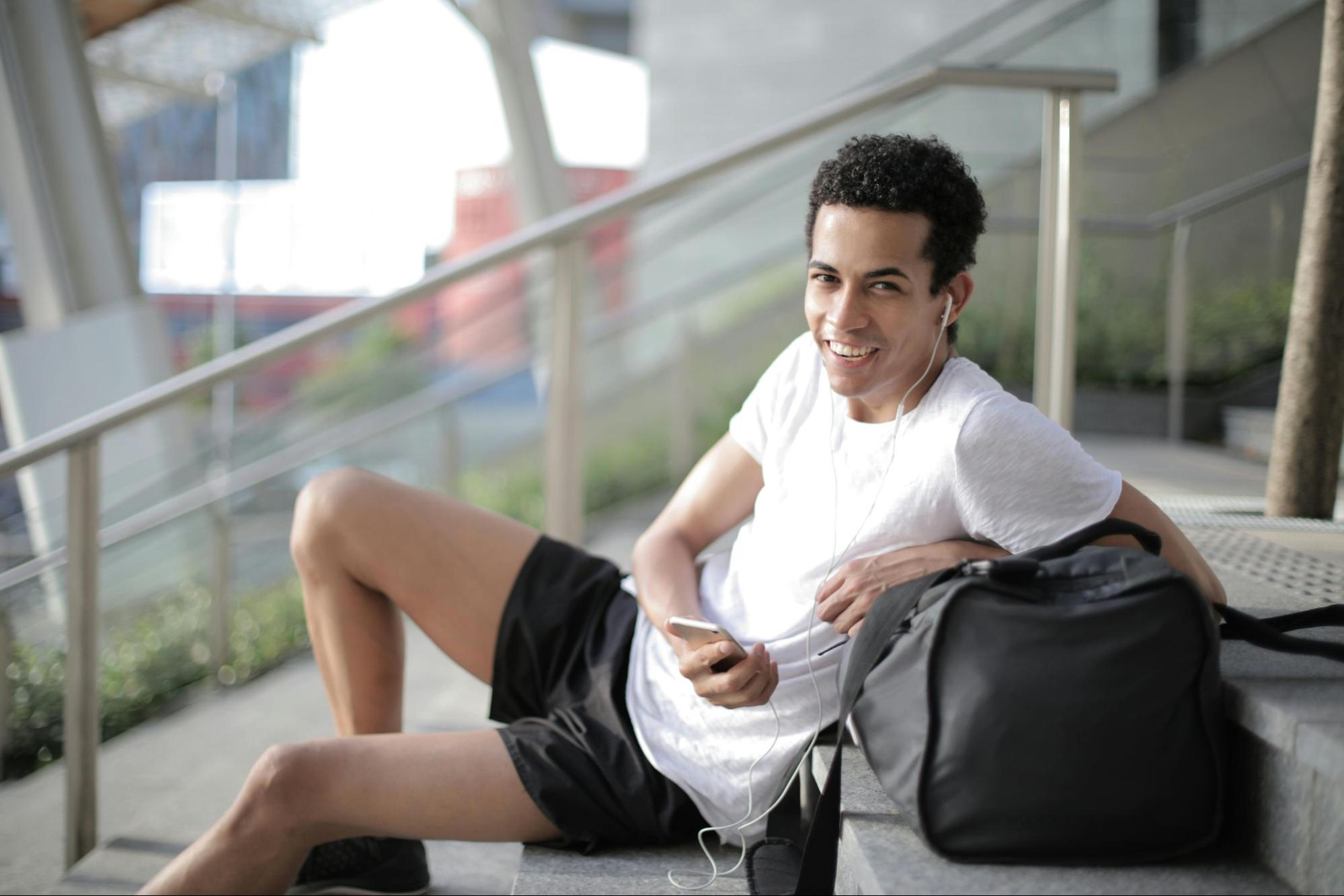 A man wearing lounge shorts and a T-shirt sits on steps with his iPod and black bag.